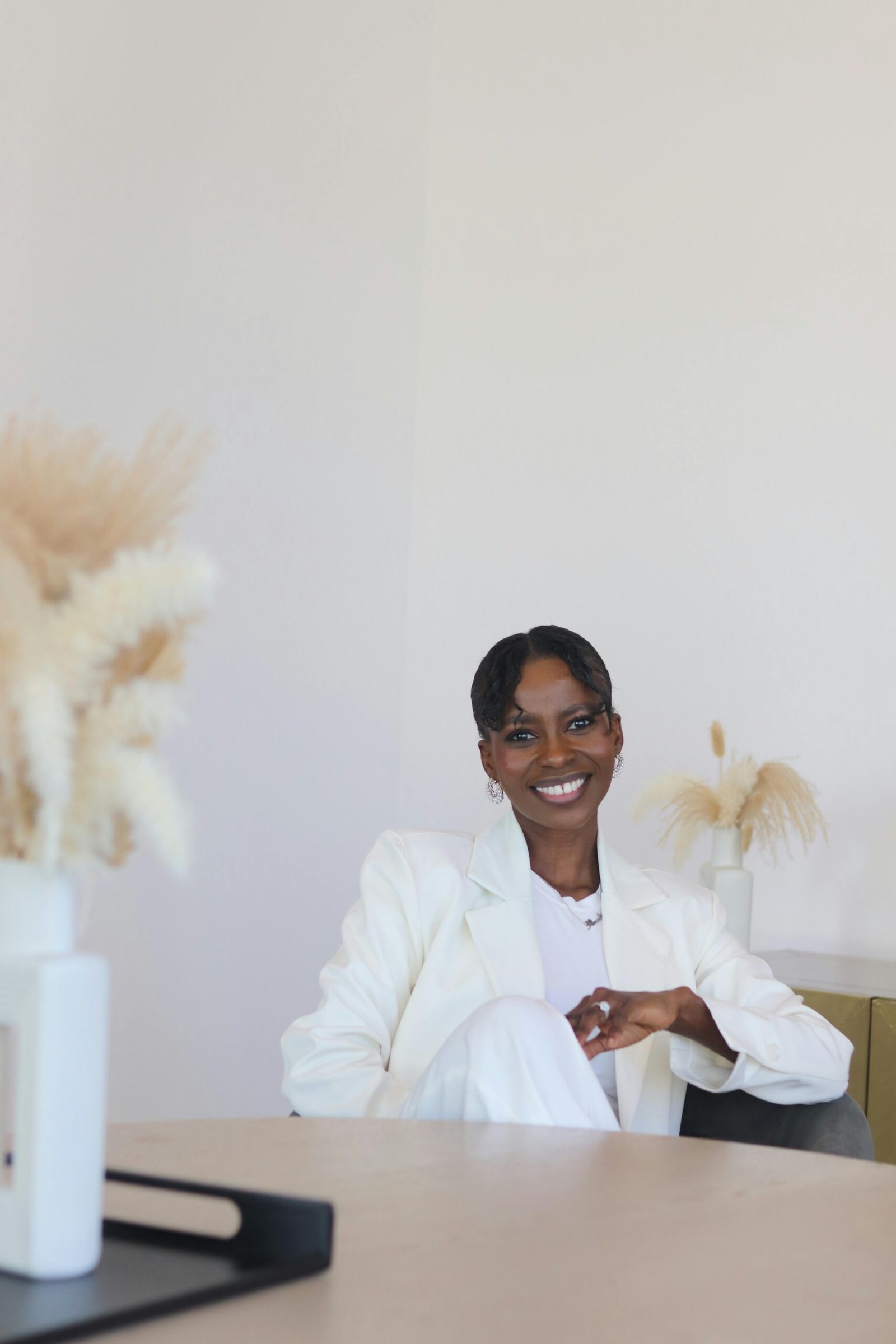 Stylish African American woman in a white suit, seated in a minimalist room with pampas grass.